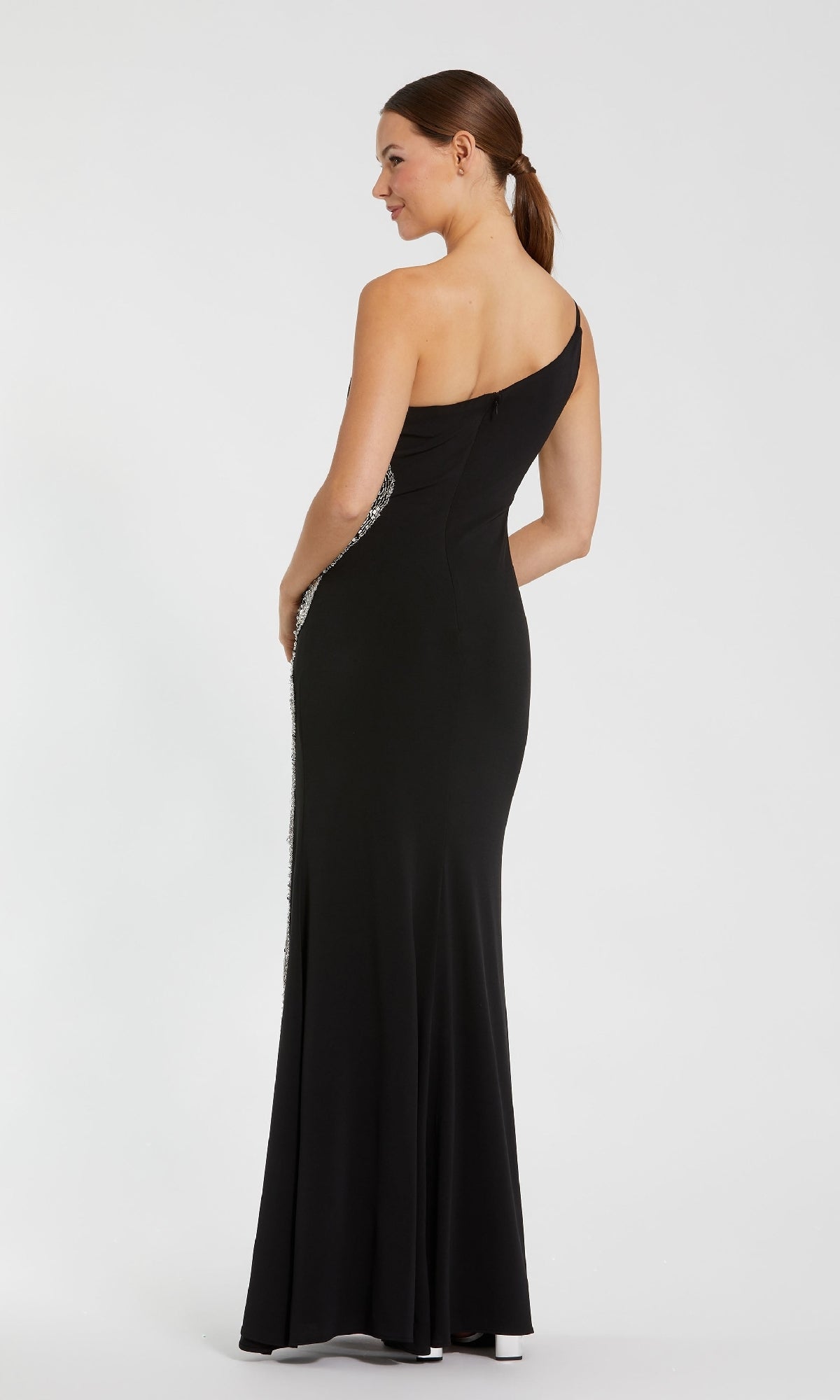 A woman models the Mac Duggal 68932 long, fitted black one-shoulder formal dress, standing with her back to the camera and looking over her shoulder against a plain white background.